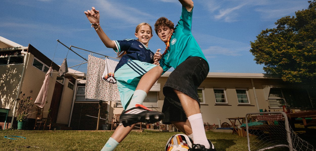 Two children playing soccer in a backyard, one kicking the ball as they both jump, with a house and laundry in the background.