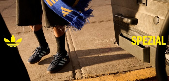 Osoba nosiaca čierne tenisky Adidas Spezial s bielymi pruhmi, čierne ponožky, krátke nohavice a drží modro-žltý šál na slnečnom chodníku.