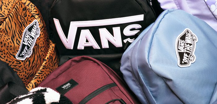 Zaini VANS in vari colori: nero con logo bianco, arancione con motivo a tigre, tessuto bordeaux e blu con accenti a contrasto.