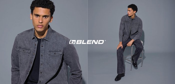 Graue Jeansjacke mit Vordertaschen, getragen über einem schwarzen Hemd. Modell sitzt auf einem Stuhl, kombiniert mit dunkler Hose und schwarzen Schuhen.