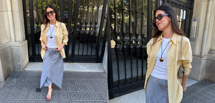 Femme souriante portant des lunettes de soleil, une chemise jaune, un haut blanc, une jupe grise et des sandales, debout dehors près d'un portail noir et d'un bâtiment en pierre.