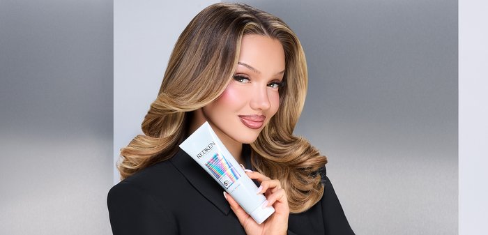 Woman with styled blonde hair holding a white Redken hair product tube, wearing a black outfit, against a gray gradient background.
