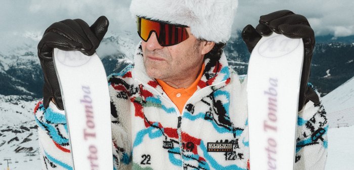 Uomo con giacca a motivi colorati, cappello di pelliccia bianca e occhiali da sci arancioni tiene in mano sci bianchi con la scritta "Roberto Tomba" sullo sfondo di una montagna innevata.