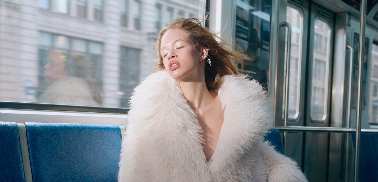 Manteau en fausse fourrure blanche avec un décolleté en V profond, une texture douce et un large col, porté sur une épaule nue, assis sur un banc de métro bleu.