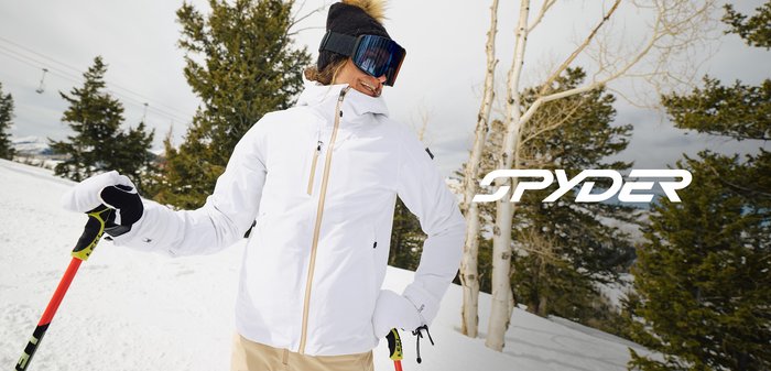 Weißer Skijacke mit hohem Kragen und Reißverschluss, akzentuiert mit einem Fell-Pompon auf einer schwarzen Mütze. Die Trägerin hält rote Skistöcke im Schnee.