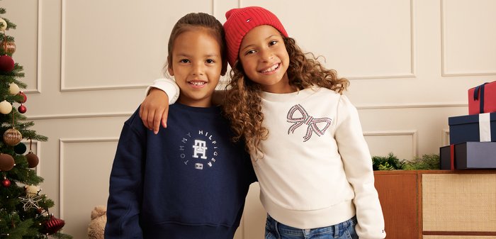Two smiling children stand close together; one wearing a navy sweatshirt and trousers, the other in a white sweatshirt with a bow design, a red beanie, and jeans.