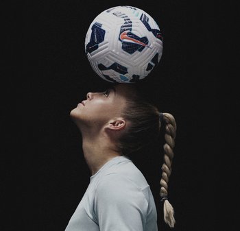 Fußball, der auf dem Kopf einer Person mit geflochtenem blonden Haar balanciert, die ein hellgraues Langarmshirt trägt, vor einem dunklen Hintergrund.
