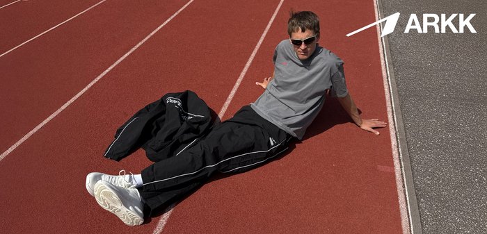 Person iført solbriller og sportsbeklædning, der læner sig tilbage på en rød løbebane ved siden af en sort jakke, ARKK-logo synligt i hjørnet.