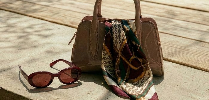 Καφέ τσάντα χειρός από δερμάτινο λουστρίνι με ένα μεταξωτό φουλάρι με σχέδια περασμένο πάνω της, δίπλα σε σκούρα κόκκινα οβάλ γυαλιά ηλίου σε ξύλινη επιφάνεια.