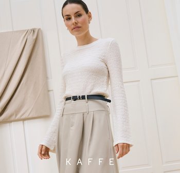 Woman wearing a white textured long-sleeve top tucked into high-waisted beige trousers with a black belt, standing against a light wall.