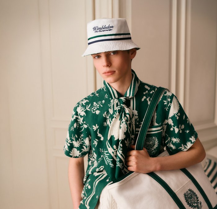 Giovane persona che indossa una camicia floreale verde, cappello a secchiello bianco Wimbledon, sciarpa coordinata e tiene una borsa da tennis bianca con rifiniture verdi.