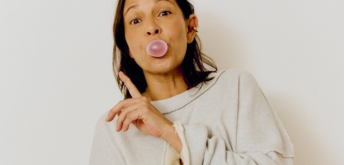 Woman wearing a light grey oversized sweatshirt and trousers blowing a pink bubble gum bubble, raising one finger in a playful gesture.