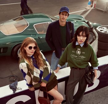 Trois mannequins posent en tenues décontractées. La femme à gauche porte un pull en argyle bleu et vert et des lunettes de soleil. L'homme au centre a une veste sombre. La femme à droite porte un sweat-shirt vert sur une chemise à col et un pantalon gris. Une voiture de sport verte est visible en arrière-plan.