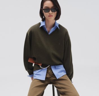 Olivengroene V-hals trui over een lichtblauw hemd, gecombineerd met beige broek, een bruine riem en donkere zonnebril. Gladde texturen.