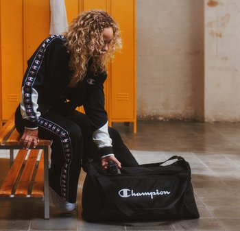 Black athletic duffel bag with white "Champion" logo. Features durable fabric, rectangular shape, and zippered opening. Minimalist design.
