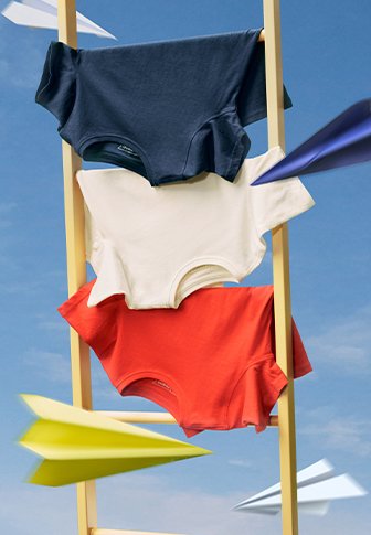 Três t-shirts de manga curta em azul marinho, branco e vermelho estão penduradas em uma escada amarela contra um céu azul, com aviões de papel nas proximidades.