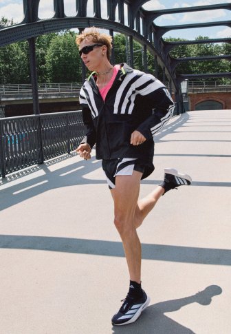 Veste de sport rayée noire et blanche avec des accents néon, t-shirt rose et shorts noirs. En train de courir sur un chemin avec une rampe en métal.