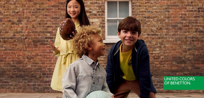 Tres niños sonrientes al aire libre junto a una pared de ladrillos, uno sosteniendo un balón de fútbol americano, otro un balón de fútbol, vestidos con ropa casual y colorida.