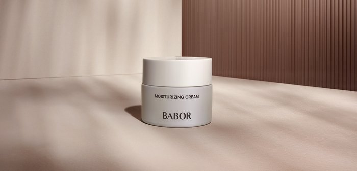 Crème hydratante dans un contenant rond en plastique blanc. Étiquette avec du texte noir sur un fond gris mat. Éclairage doux, surface lisse.