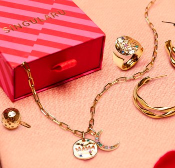 Collana a catena dorata con ciondoli a forma di mezzaluna con pietre preziose e la scritta "MAMA", accanto a un anello d'oro decorato con gemme, orecchini a cerchio e una scatola rossa a strisce.