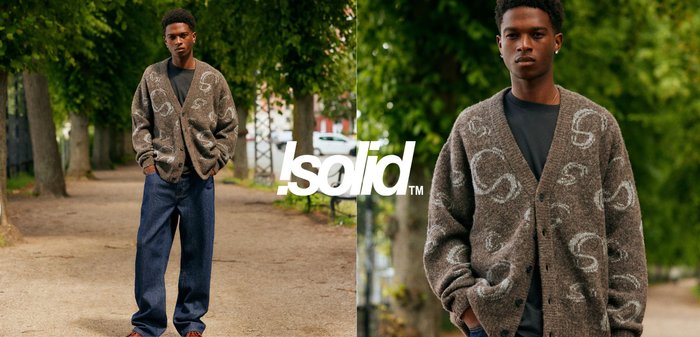 Braun gemusterter Cardigan mit V-Ausschnitt, strukturiertem Strick und Knöpfen, kombiniert mit dunkelblauen, locker sitzenden Jeans. Grünes Laub im Hintergrund.