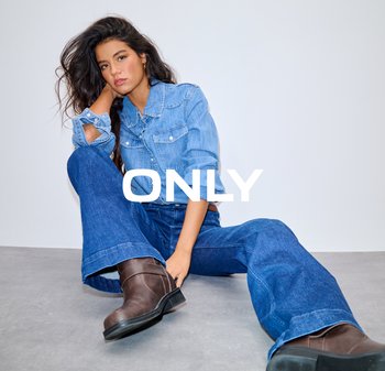 Jonge vrouw met lang donker haar, gekleed in een denim blouse, wijd uitlopende spijkerbroek en bruine laarzen, zittend op een grijze vloer tegen een witte muur.