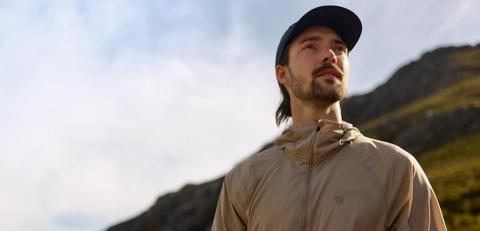 Man wearing a beige jacket and black cap looking upward outdoors with rocky hills and cloudy sky in the background.