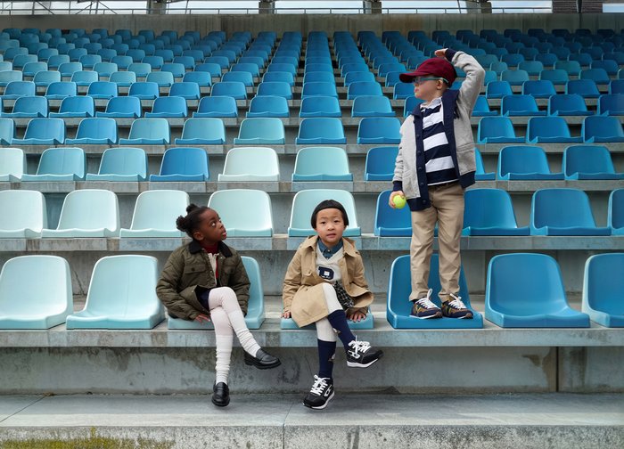 Trois enfants sont assis sur des gradins de stade. Les sièges sont bleu pâle. Chaque enfant porte une tenue distincte : une veste verte, un trench-coat beige et un sweat à capuche gris.