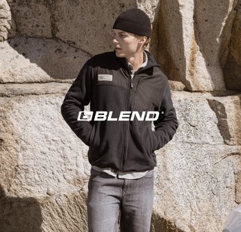 Nero giacca in pile con una tasca sul petto, camicia grigia sottostante, abbinata a jeans grigi chiari e un berretto nero. Logo in testo visibile.