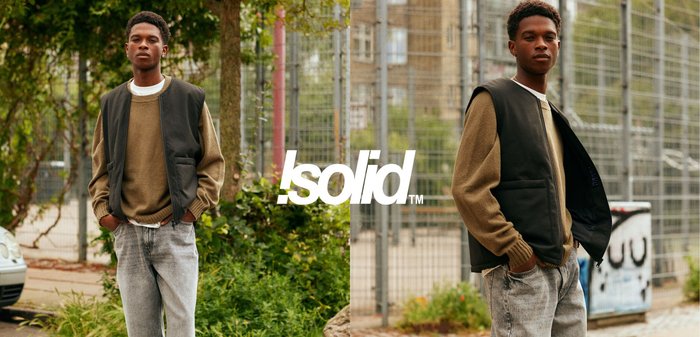 Ein junger Mann mit braunem Pullover und schwarzer Weste steht lässig im Freien nahe Grünflächen und einem Metallzaun in einer städtischen Umgebung, die Hände in den Taschen.