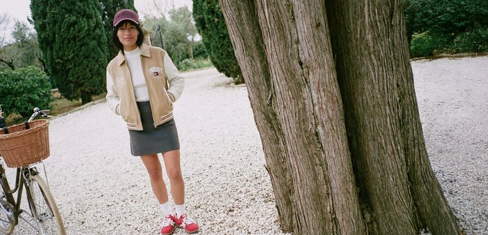 Jeune femme portant une casquette, une veste beige et une jupe, debout sur un chemin de gravier à côté d'un grand arbre avec un vélo à proximité dans un parc.