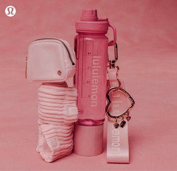 Borraccia rosa Lululemon con segni di misurazione, elastici bianchi impilati, pochette piccola e portachiavi a forma di cuore su sfondo rosa.