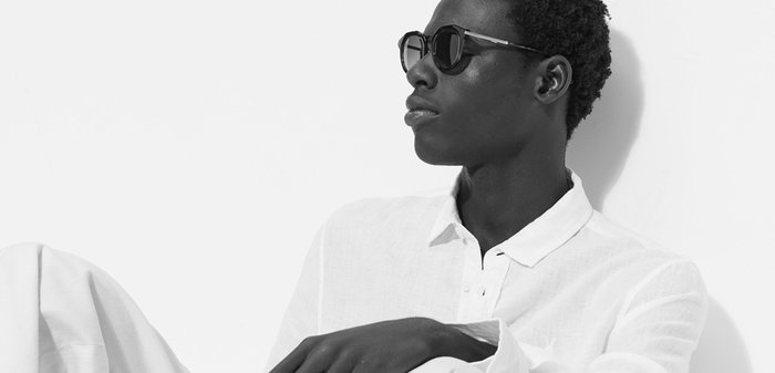 Homme portant des lunettes de soleil et une chemise blanche ample avec un pantalon blanc, assis en position détendue contre un mur blanc dans une lumière vive et minimaliste.
