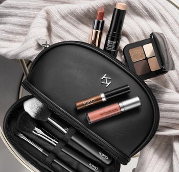 Black cosmetic bag with silver logo, containing brushes and makeup items: lipsticks, highlighting stick, eyeshadow palette with neutral tones.