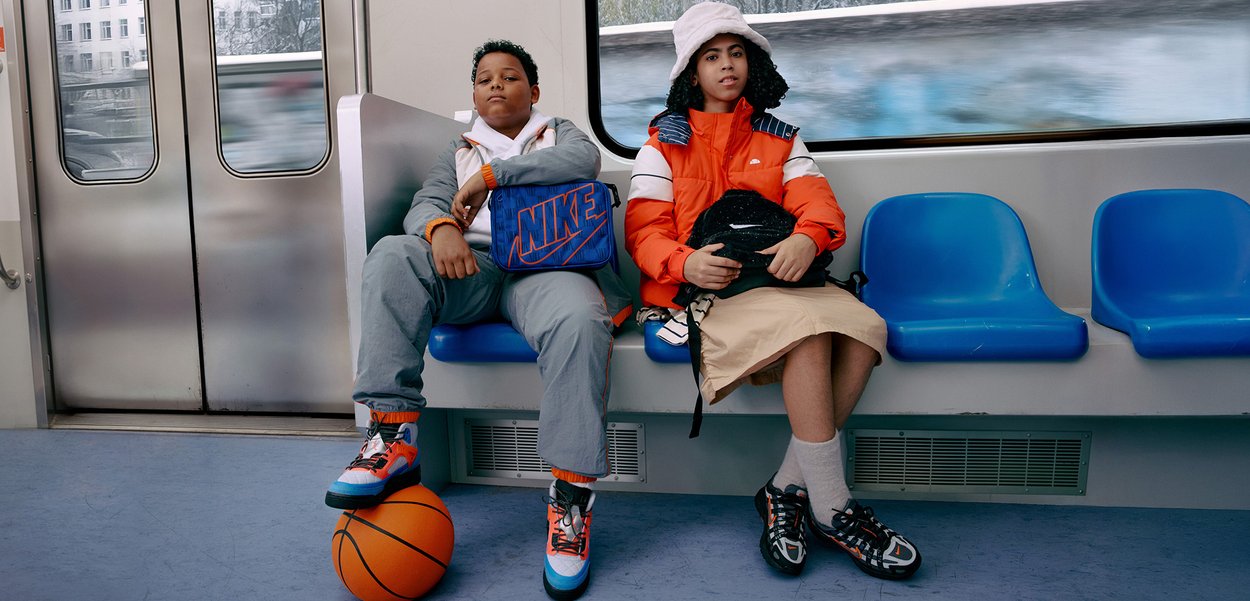 Två personer sitter på en tunnelbanebänk. Den första bär en grå träningsdräkt och en blå Nike-väska, och den andra har en orange dunjacka och kjol.