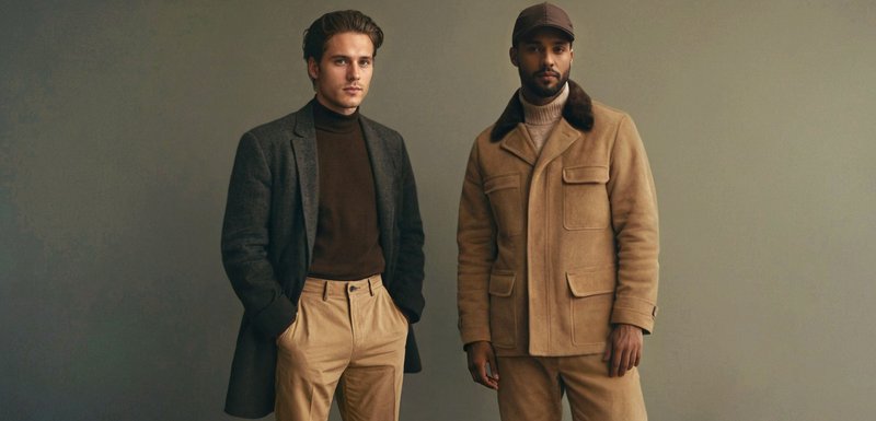 Deux hommes portant des vêtements d'automne : l'un en manteau gris foncé avec un col roulé marron et un pantalon beige, l'autre en veste beige, col roulé, casquette et pantalon.