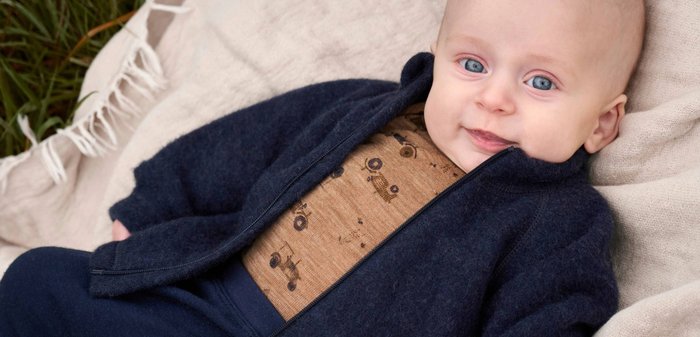 Brauner gestrickter Cardigan über einem hellbraunen Overall mit schwarzem Tiermuster. Baby auf einer weichen Oberfläche, mit zwei Birnen neben sich. Helle Texturen und warme Farbtöne.