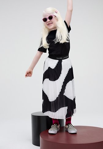 Μαύρη μπλούζα, πλισέ φούστα με μαύρο-άσπρο σχέδιο, ροζ κάλτσες, μαύρα-άσπρα καρό αθλητικά παπούτσια και στρογγυλά γυαλιά ηλίου.