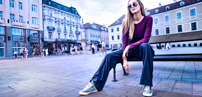 Junge Frau mit Sonnenbrille, lila langärmeligem Oberteil und lockeren Hosen, die auf einer Bank auf einem Stadtplatz mit historischen Gebäuden sitzt.