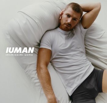 Uomo che indossa una maglietta bianca e pantaloni corti neri sdraiato su un letto bianco con un braccio dietro la testa, il marchio "IUMAN" visibile.