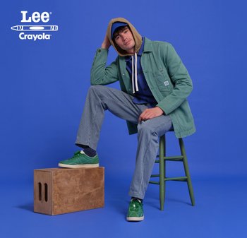 Veste en velours côtelé vert sur un sweat à capuche bleu, pantalon rayé et baskets vertes. Caisse en bois avec des poignées découpées au premier plan, fond bleu.