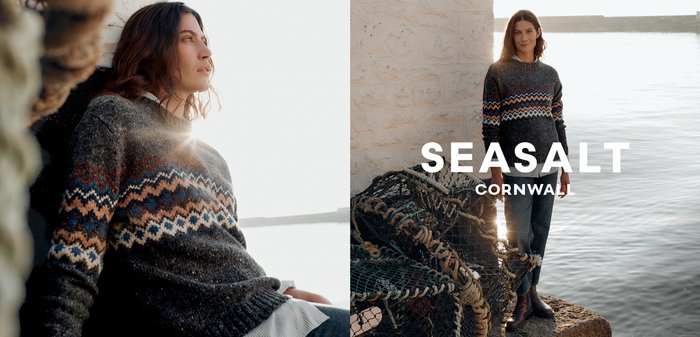 Šedý vlněný svetr s geometrickými vzory v modré, hnědé a bílé barvě. V kombinaci s džíny a hnědými botami, na pozadí pobřežní scenérie.