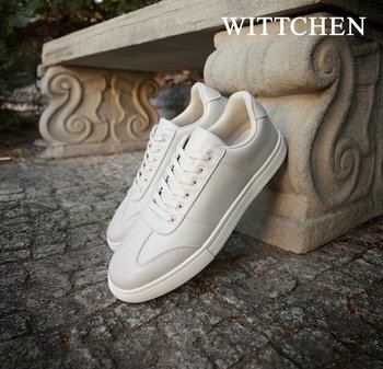 Białe skórzane sneakersy o gładkiej fakturze, sznurowanym designie i wzorzystych gumowych podeszwach. Umieszczone blisko kamiennej ławki.