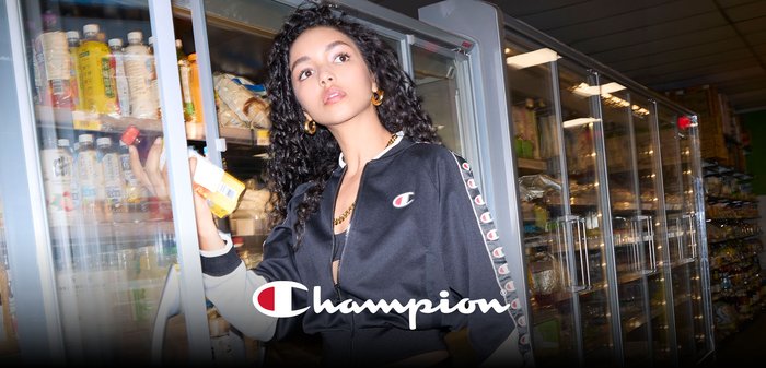 Black zip-up track jacket with white and red accents, worn over a black top, held by a person in front of a refrigerator stock.