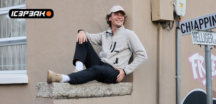 Un homme en veste gris clair, pantalon noir et casquette blanche est assis en souriant sur un rebord en pierre près de panneaux de signalisation et d'un mur beige.