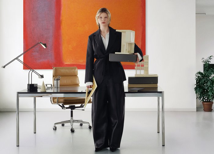Γυναίκα με μαύρο κοστούμι κρατώντας αρχιτεκτονικά μοντέλα και μια καρτέλα, στέκεται σε μοντέρνο γραφείο με γραφείο, καρέκλα, λάμπα και πορτοκαλί αφηρημένο πίνακα.