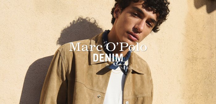 Młody mężczyzna w beżowej zamszowej kurtce i białej koszuli, stojący przy beżowej ścianie, na którą pada cień jego postaci.