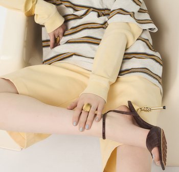 Striped grey and mustard t-shirt, soft yellow shorts, brown textured sandals, and a large gold ring on a hand with pastel nails.