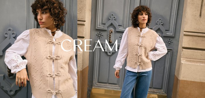 Beige gestrickte Weste mit strukturiertem Muster, Zopfmuster und dekorativen Schleifenverschlüssen, kombiniert mit einer weißen Bluse mit Lochstickerei an den Manschetten.