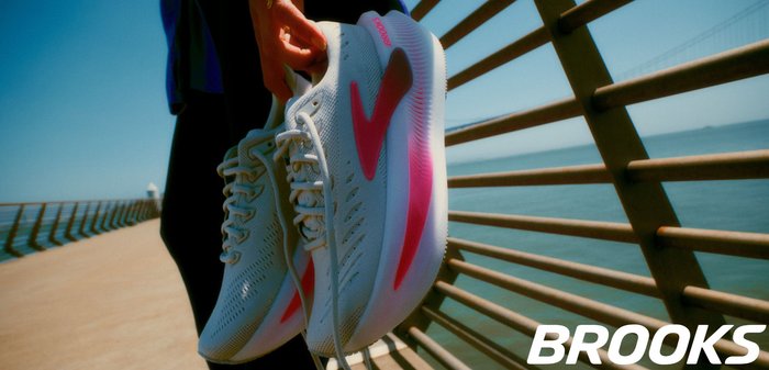 Scarpe da corsa bianche con accenti rosa, parte superiore in rete testurizzata e design snello. Il marchio "Brooks" è visibile sul lato della scarpa.